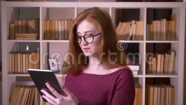 在大学图书馆里用平板电脑拍摄的戴眼镜的年轻红发美女白种人的特写照片